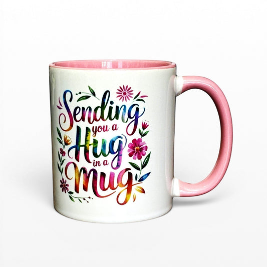 White ceramic mug with pink handle and interior, featuring colourful "Sending you a Hug in a Mug" text and floral design.