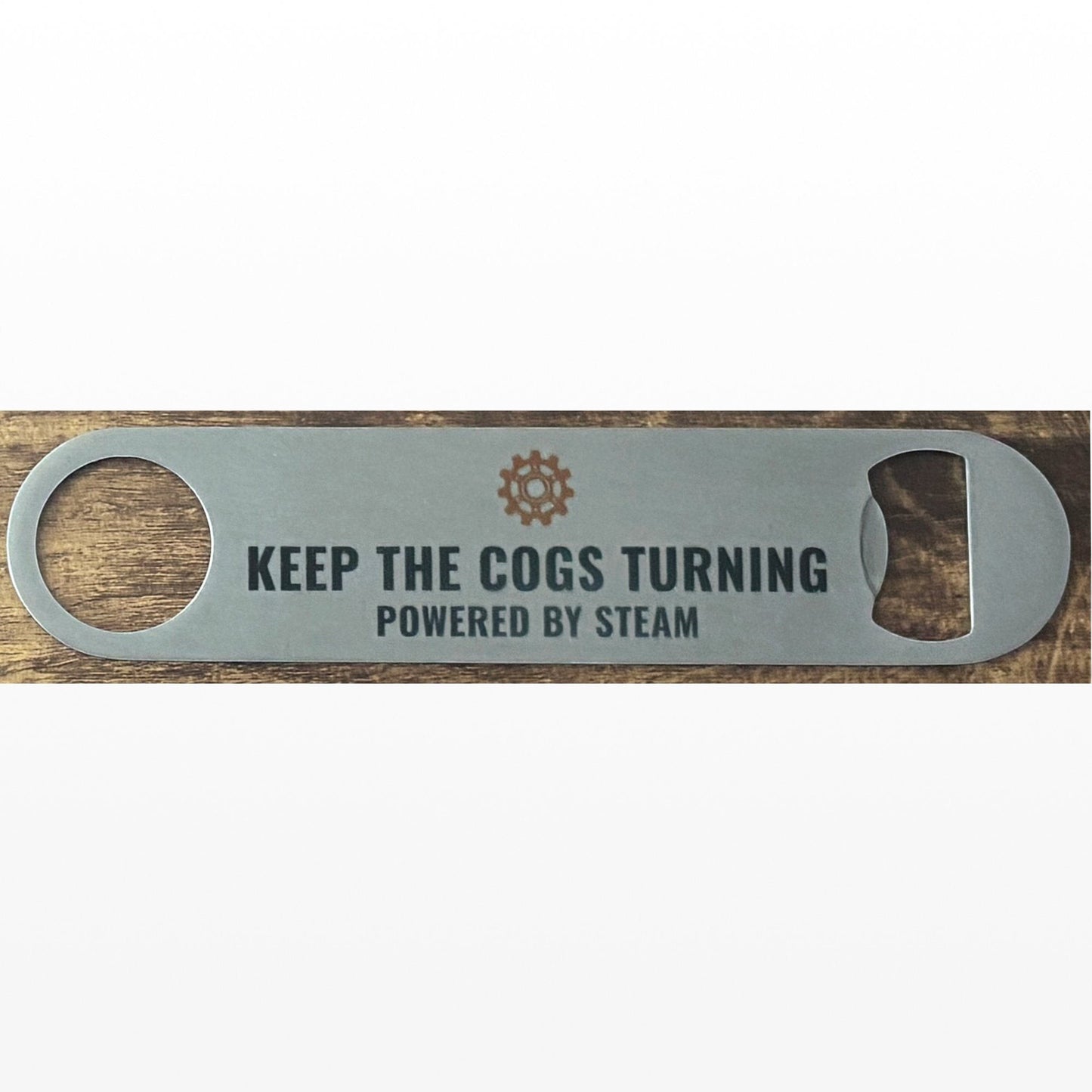 Stainless Steel Bar Blade with Steampunk Cog and Keep The Cogs Turning Text in black. Bar blade on wooden table.