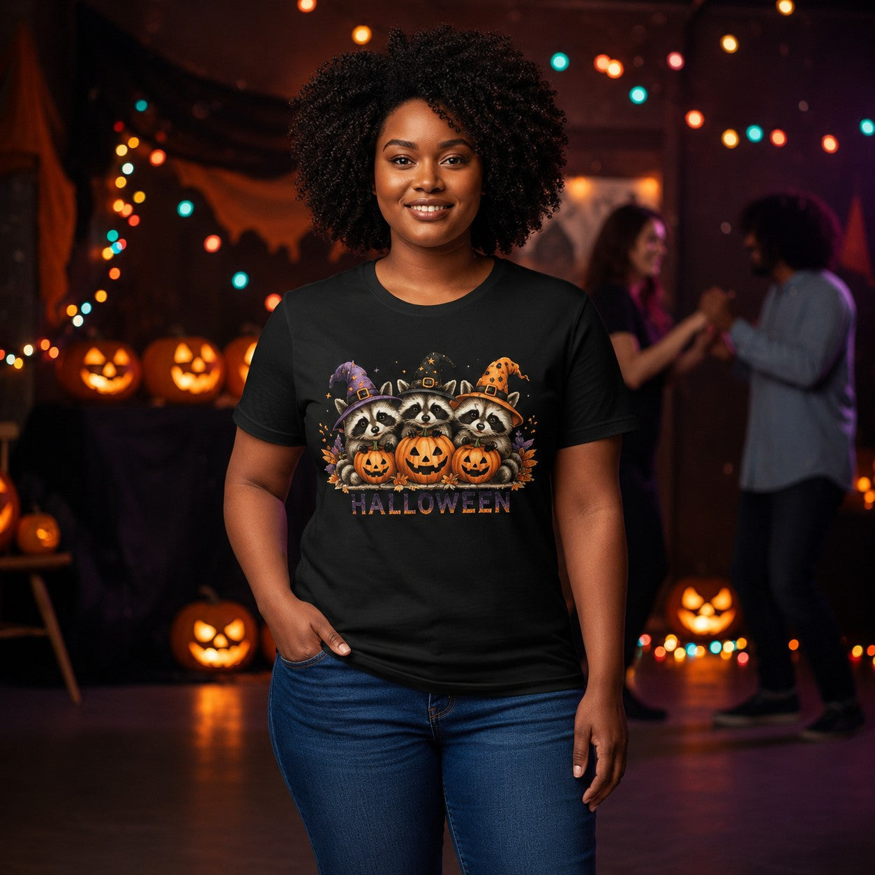 A woman wearing a black graphic t-shirt with a Halloween-themed raccoon and pumpkin design, standing in a festive indoor setting with string lights and jack-o'-lanterns in the background.
