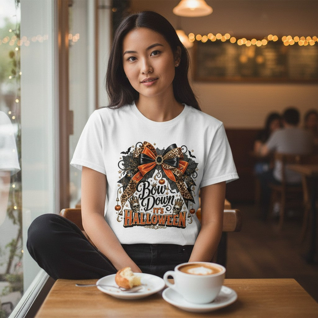 A woman sitting at a cafe table wearing a white graphic t-shirt with a Halloween design that reads 'Bow Down Halloween' with a large orange and black bow. A cup of coffee and a pastry are on the table in front of her.