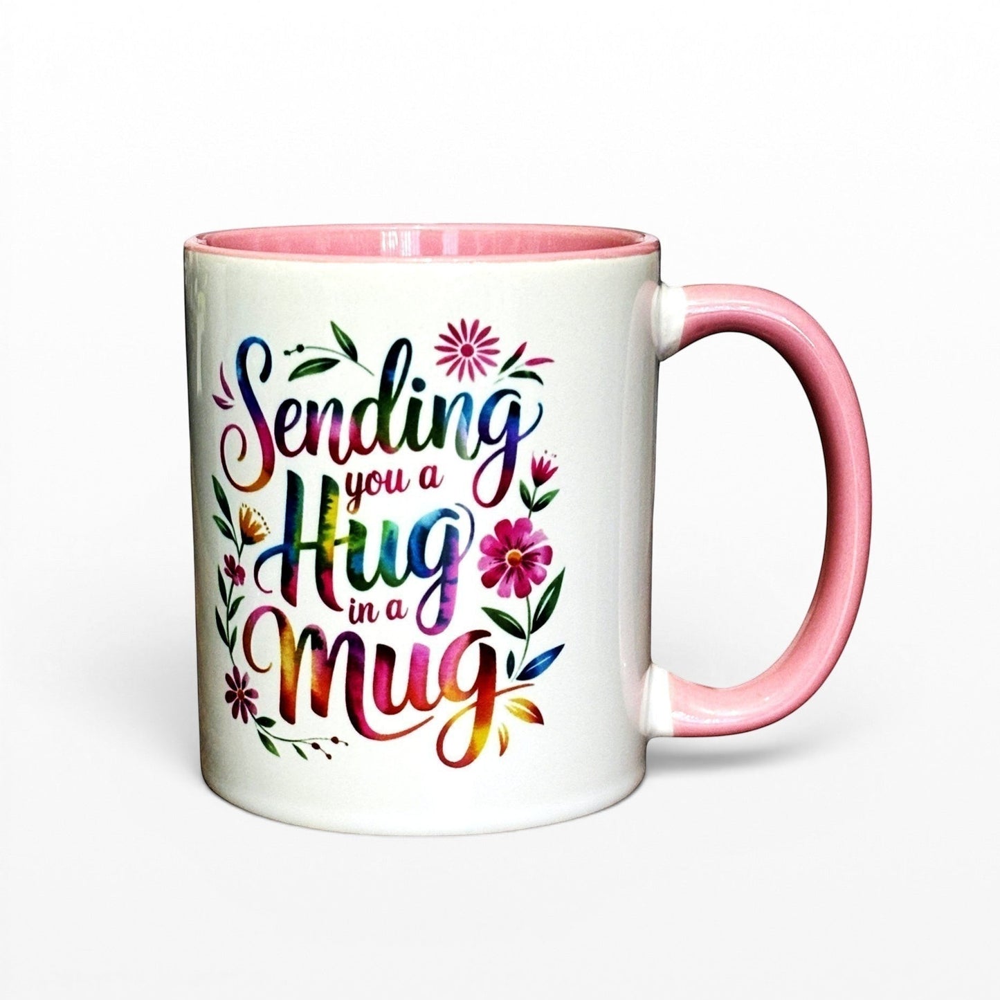 White ceramic mug with pink handle and interior, featuring colourful "Sending you a Hug in a Mug" text and floral design.