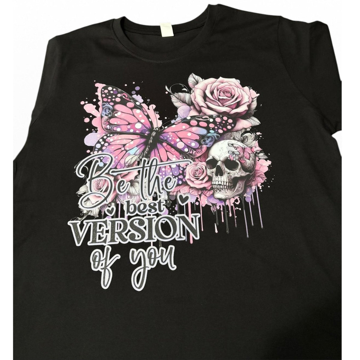 A black short-sleeve t-shirt featuring a gothic-style graphic print with a pink butterfly, pink roses, a skull, and the text 'Be the best VERSION of you' in white and black lettering.