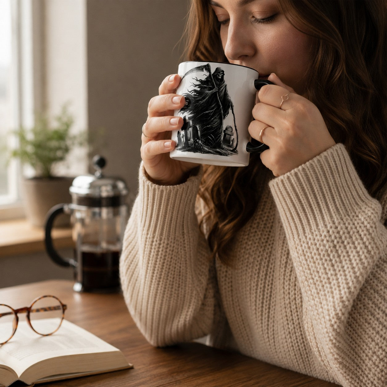 A woman in a beige knit sweater holds a white ceramic mug with a black illustration of a grim reaper to her lips in a cozy, sunlit room. A book with glasses rests on the wooden table in the foreground.