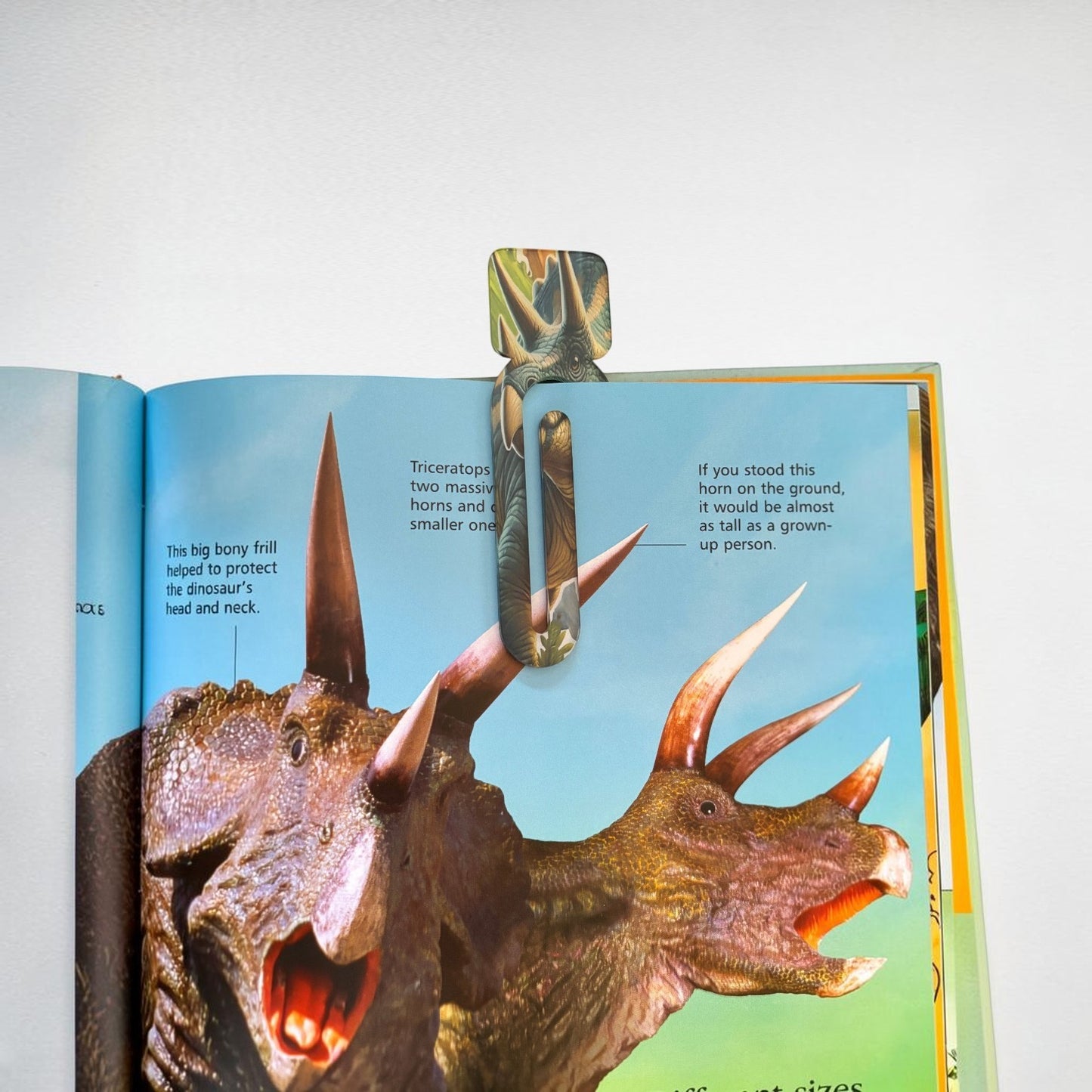 Open book with a dinosaur illustration and text on a white background, showing a book clip on the page