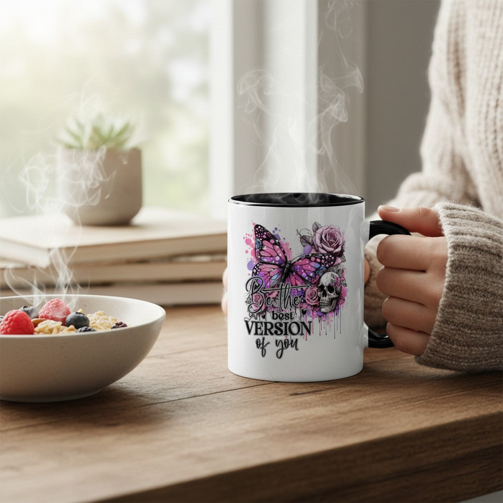 A white ceramic mug with a black handle and interior, featuring a purple and pink butterfly, skull, and rose graphic with the text 'the best VERSION of you', held by a person in a beige sweater at a wooden table next to a bowl of cereal.
