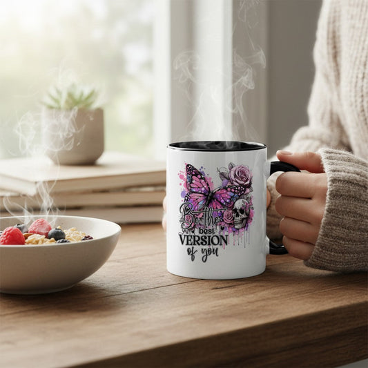 A white ceramic mug with a black handle and interior, featuring a purple and pink butterfly, skull, and rose graphic with the text 'the best VERSION of you', held by a person in a beige sweater at a wooden table next to a bowl of cereal.