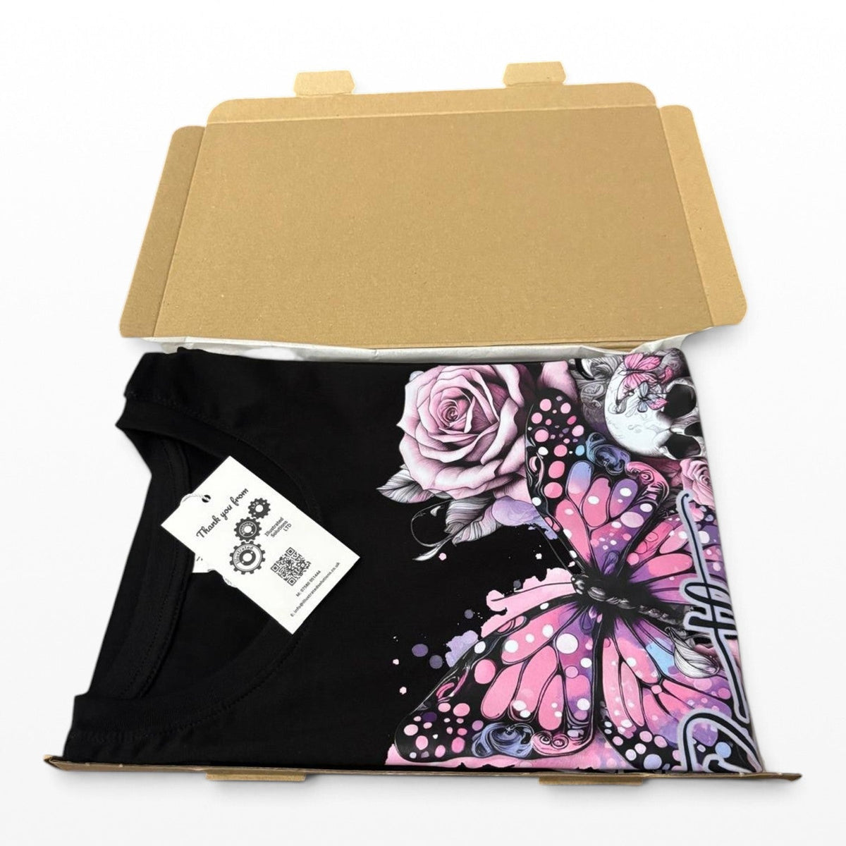 A black short-sleeve T-shirt with a large pink, purple, and white butterfly and rose graphic print, folded inside an open brown cardboard box with a white product tag attached.