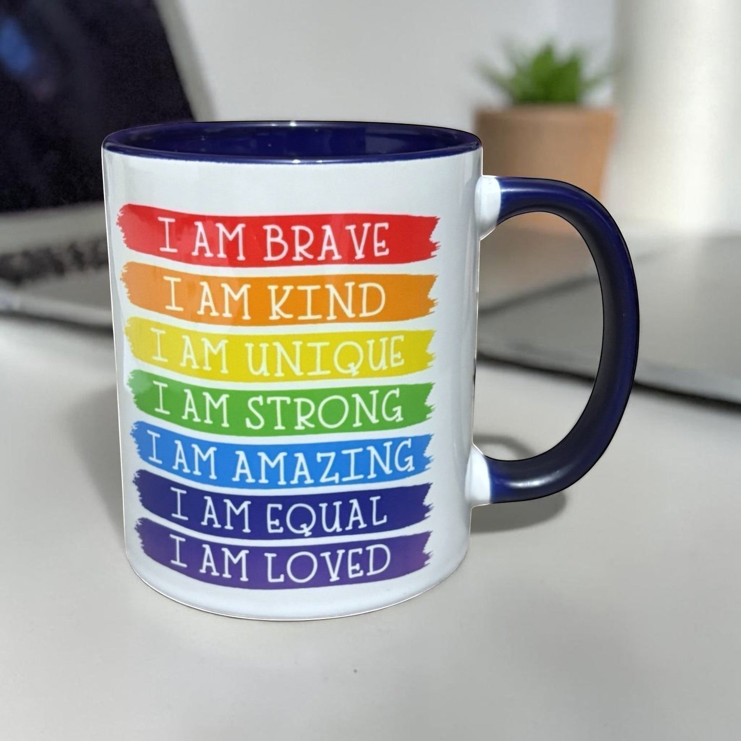 Illustrated Solutions Store white ceramic mug with navy blue interior and handle, featuring rainbow stripes with affirmations like "I Am Brave" and "I Am Loved".