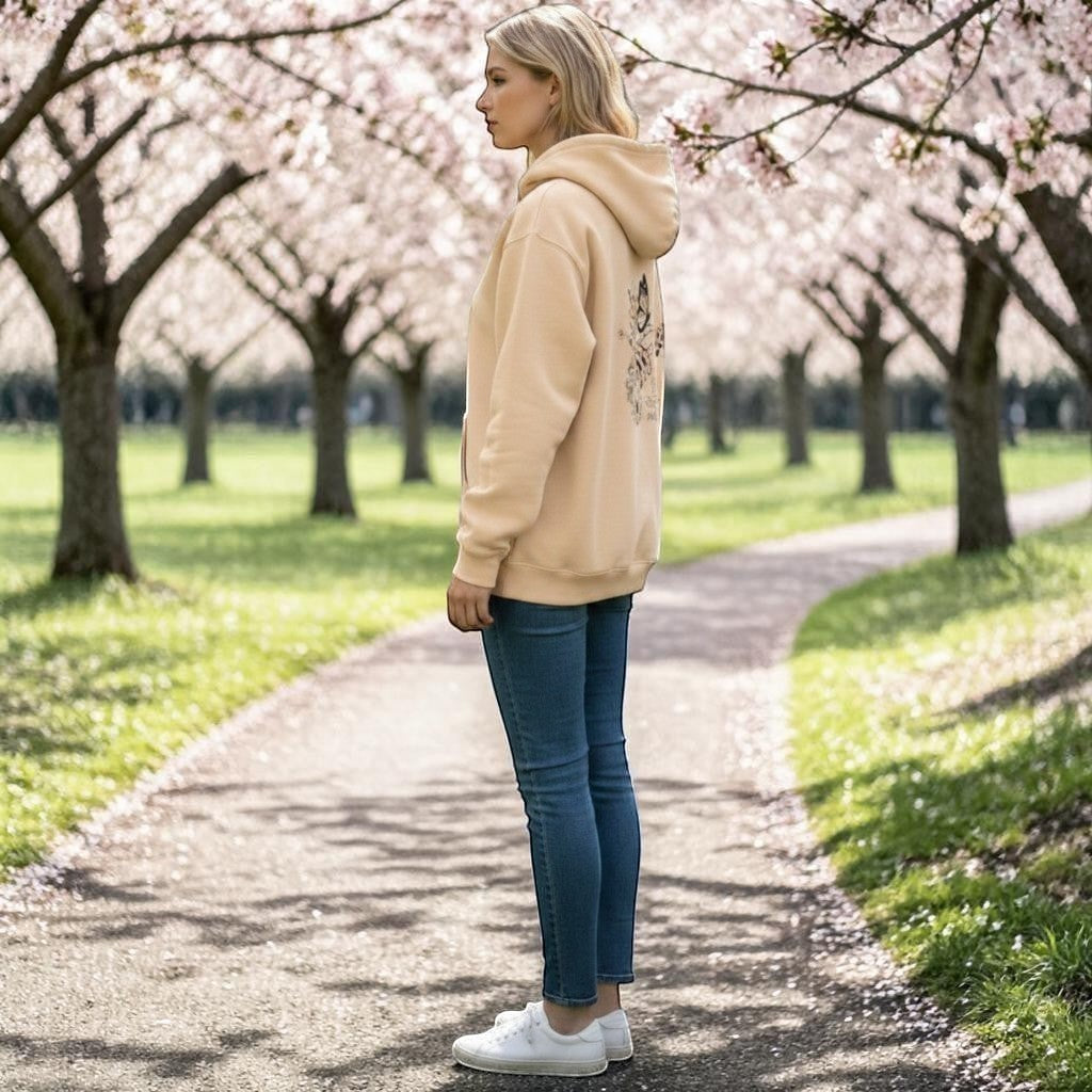 Person wearing a beige hoodie with floral designs and text, standing side on, on a path in a park.