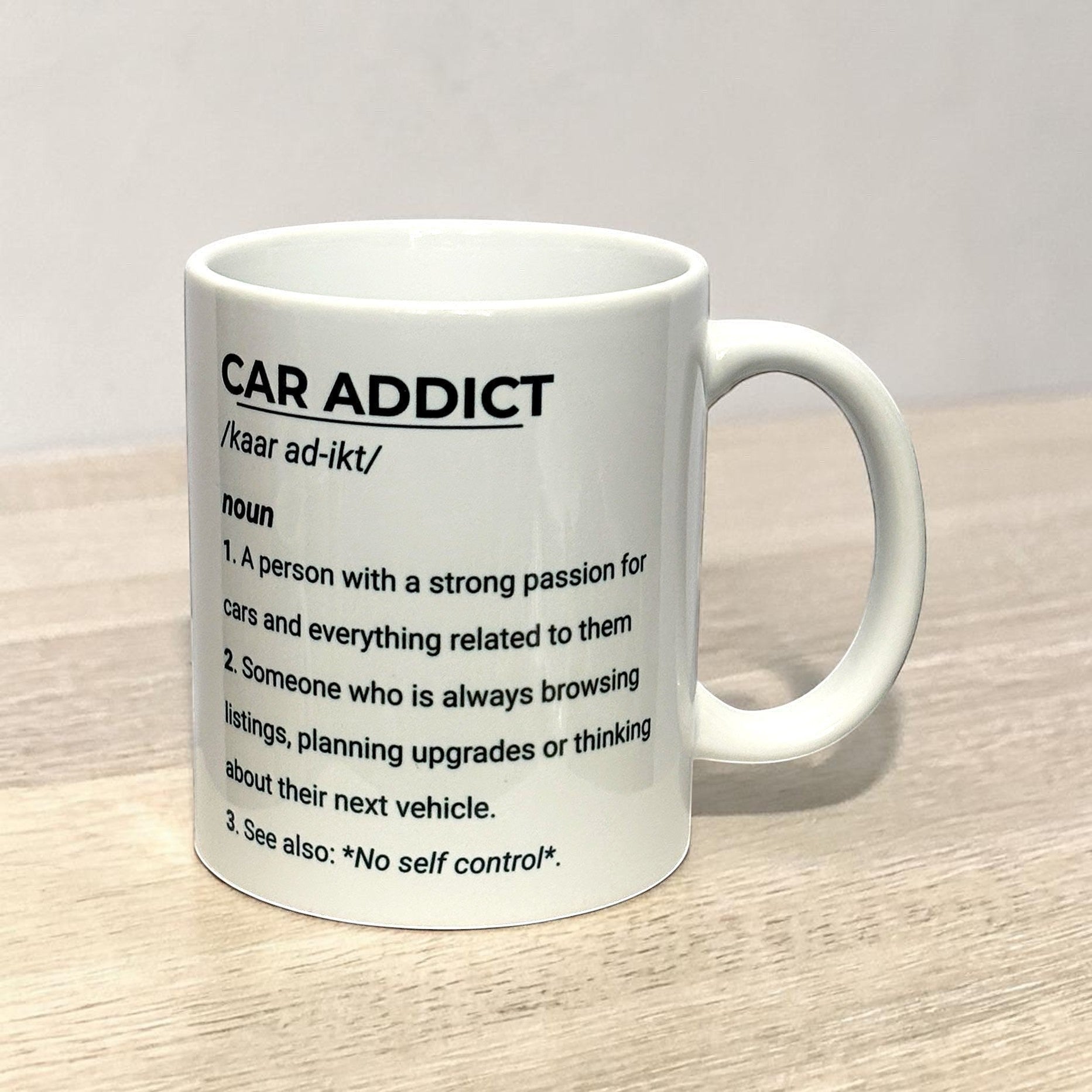Illustrated Solutions Store white ceramic mug with black text defining 'Car Addict' as a noun with a humorous definition.