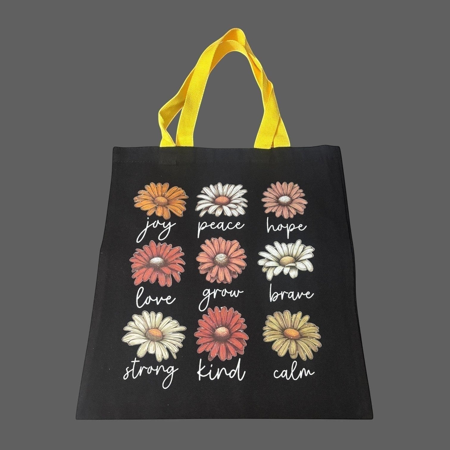Illustrated Solutions Store black canvas tote bag with yellow handles, featuring a grid of daisy illustrations with positive affirmations like joy, peace, hope, love, grow, brave, strong, kind, and calm.