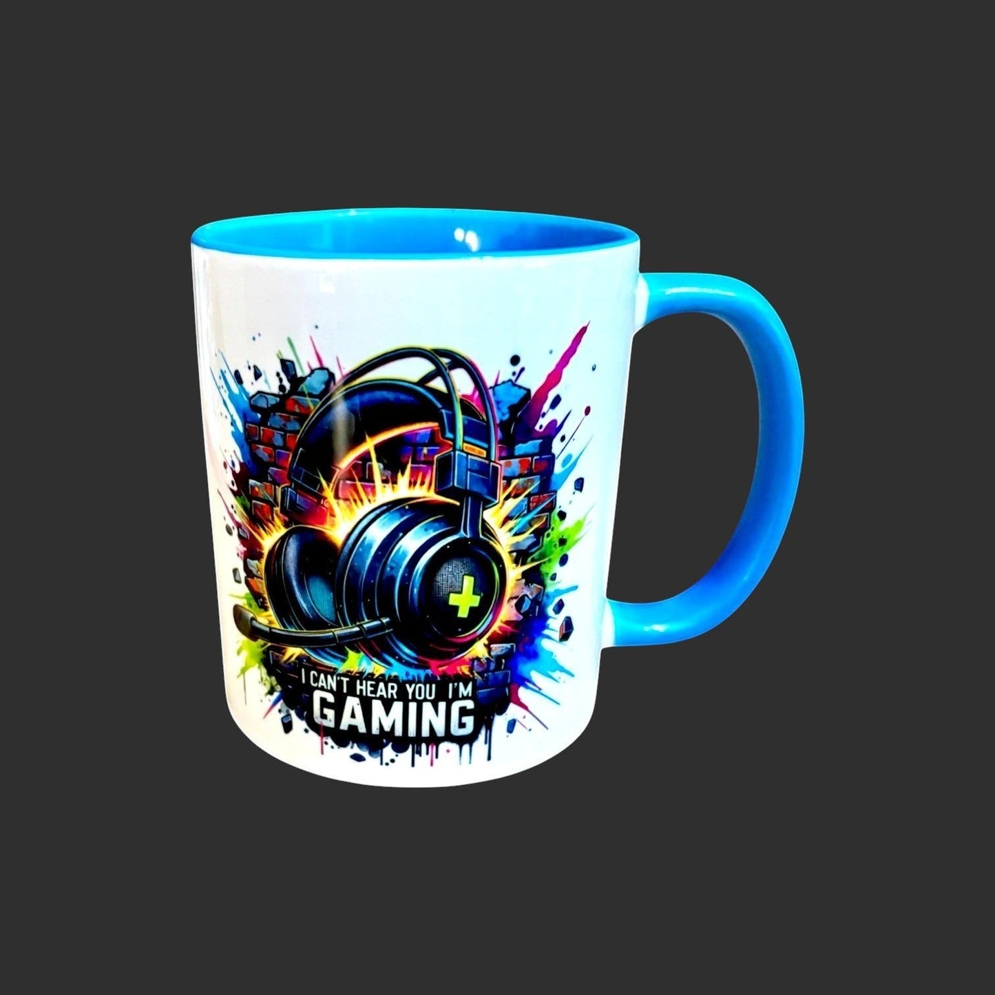 Illustrated Solutions Store white ceramic mug with blue handle and interior, featuring a graphic of black gaming headphones against a brick wall with colorful paint splatters and the text "I CAN'T HEAR YOU I'M GAMING".
