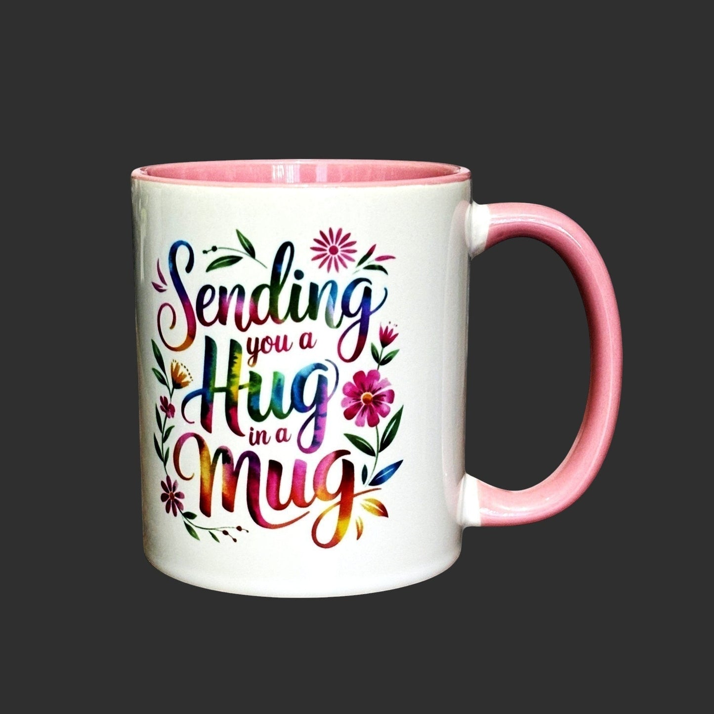 White ceramic mug with pink handle and interior, featuring colourful "Sending you a Hug in a Mug" text and floral accents. Mug on white background.