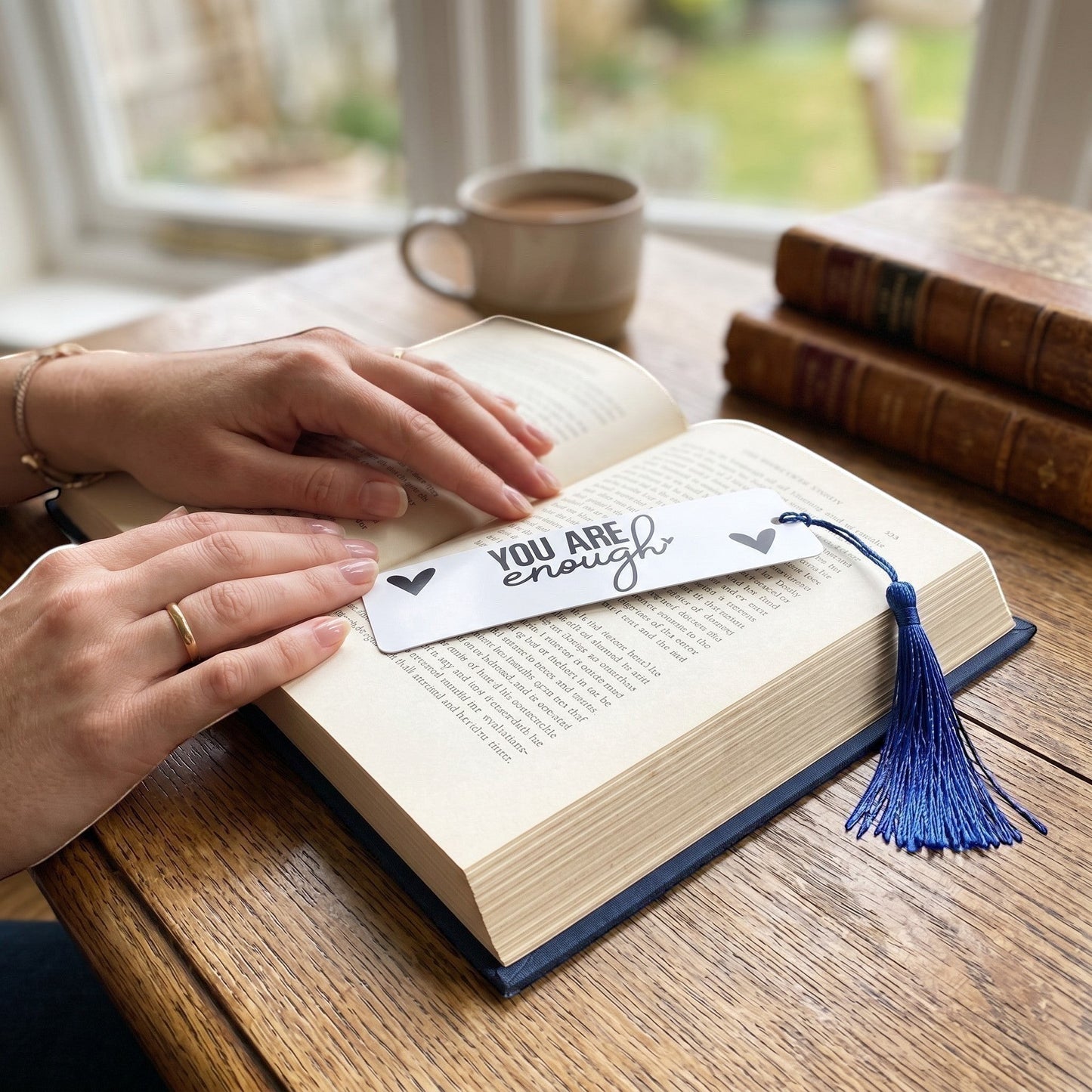 Illustrated Solutions Store metal bookmark with You Are Enough text and blue tassel resting on an open book.