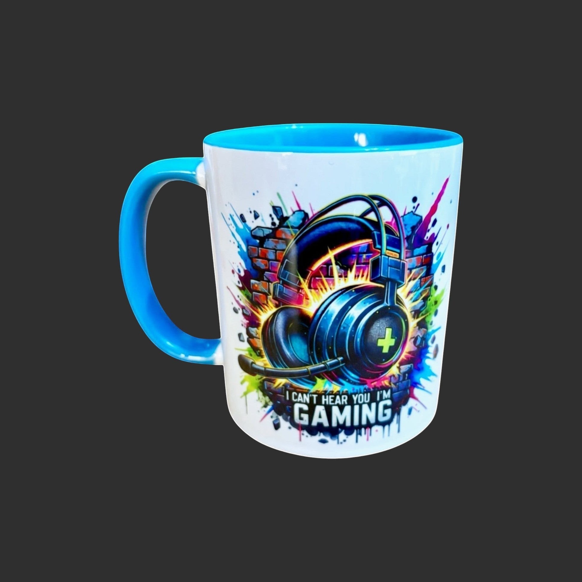 Illustrated Solutions Store white ceramic mug with blue handle and interior, featuring a graphic of gaming headphones against a brick wall with colorful paint splatters and the text "I CAN'T HEAR YOU I'M GAMING".