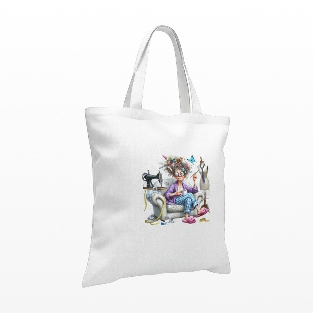 Illustrated Solutions Store white tote bag with a humorous sewing design of a woman with messy hair sitting in a chair with a sewing machine and mannequin.