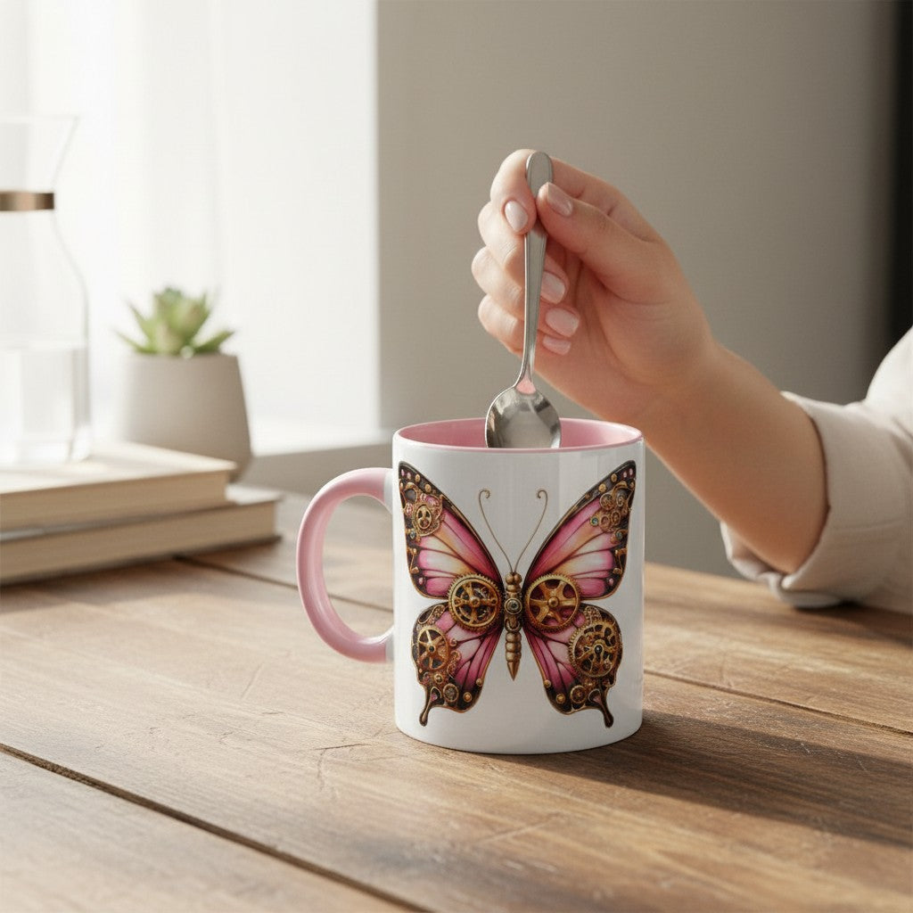 A white ceramic mug with a pink handle and interior, decorated with a large pink and gold butterfly design, sits on a wooden table. A hand holding a spoon stirs the mug, with a softly blurred indoor background.