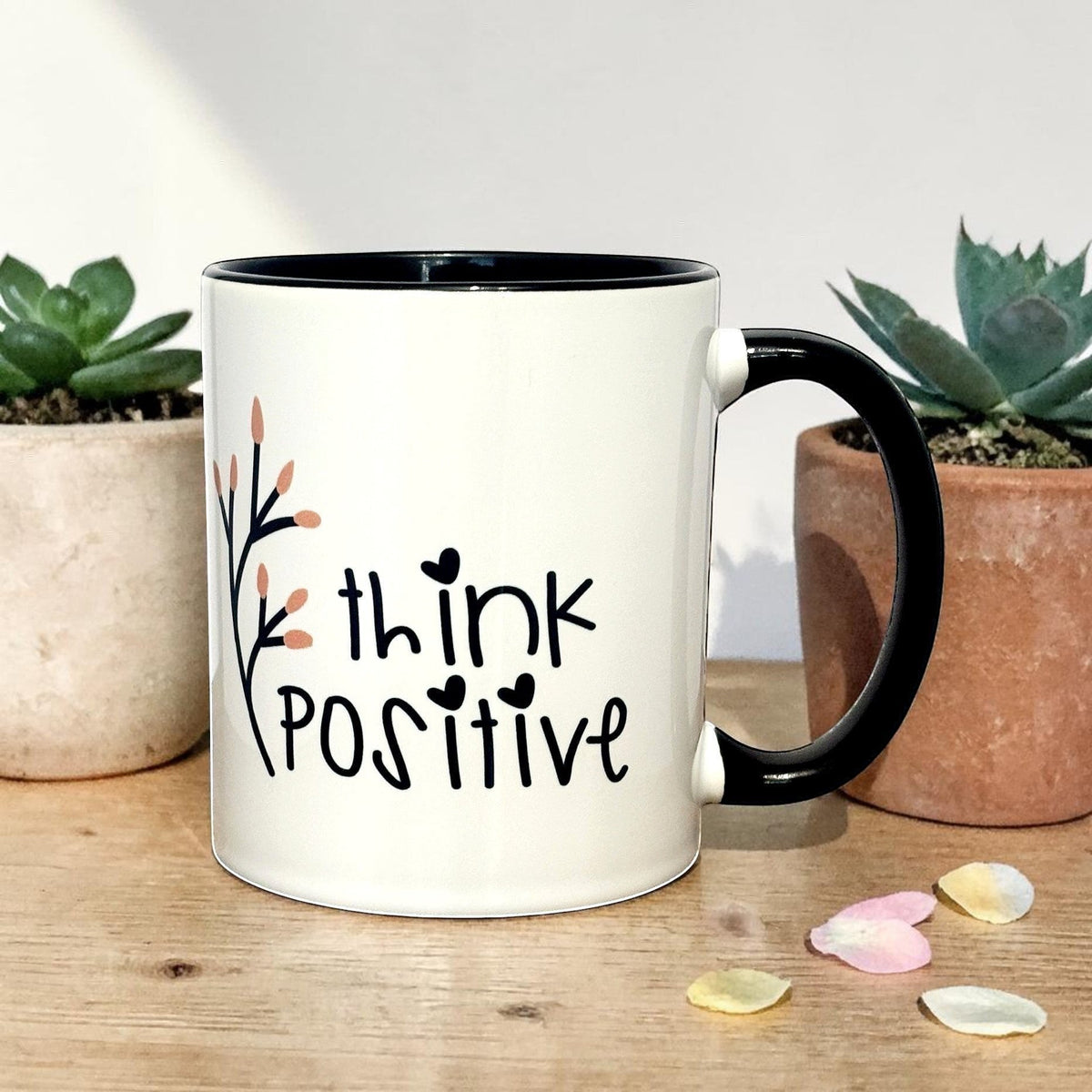 White ceramic mug with black handle and rim, featuring "think POSITIVE" text and a floral branch design. Mug placed on a table.