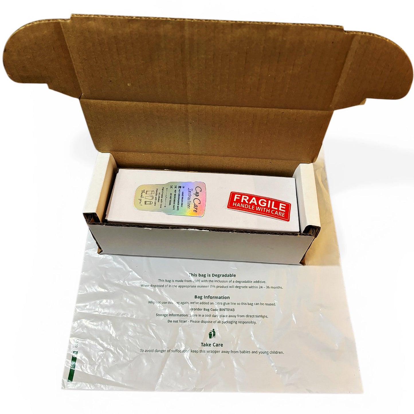 An open brown cardboard shipping box containing a white rectangular package with a rainbow holographic sticker and a red 'FRAGILE HANDLE WITH CARE' label, placed over a clear plastic degradable bag with printed handling instructions on a white background.