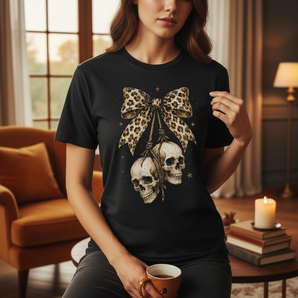 A woman wearing a black graphic t-shirt with a large beige and black leopard-print bow and two hanging skulls, posed in a cozy, warmly lit living room.
